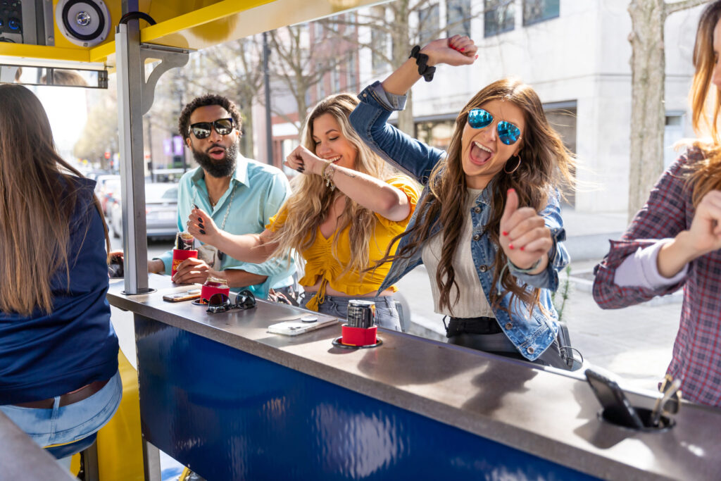 Group of friends enjoying an outdoor party, with a woman in sunglasses smiling and reaching out, and others holding drinks.