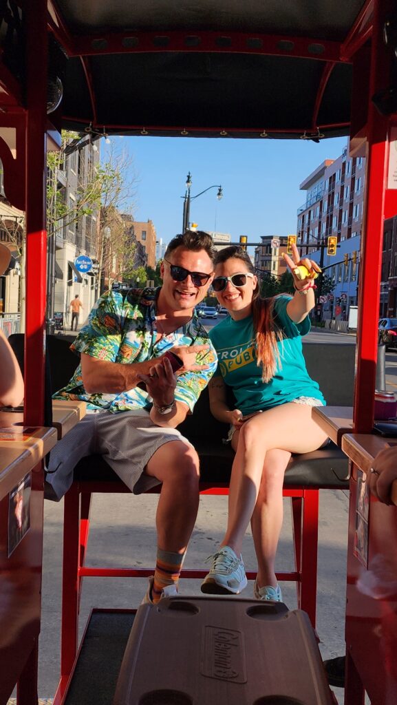 Two people smiling and posing for a photo on a city street, one showing a peace sign.