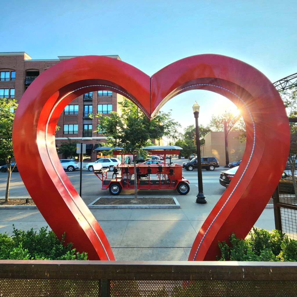 Large red heart sculpture in urban setting with sun shining through, surrounded by buildings and a unique multi-seat bicycle in the background.