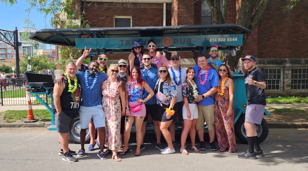 Group of people wearing tropical outfits and leis standing together in front of a trolley bus on a sunny day.