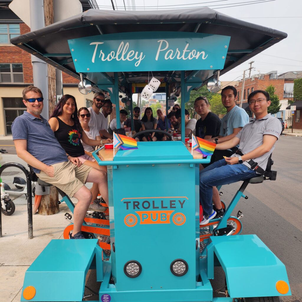 Group of people enjoying a ride on a turquoise trolley pub with rainbow flags, titled "Trolley Parton," on a city street.