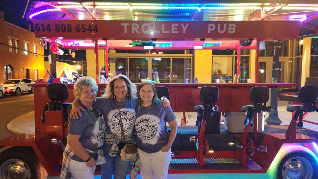 Three women posing in front of a brightly lit trolley pub at night, wearing matching "Warning: Girl Trip in Progress" shirts.