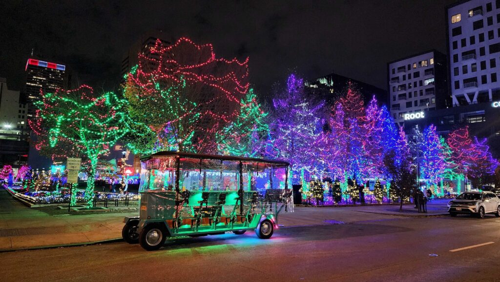 Festive city street scene with vibrant holiday lights on trees and buildings, with a decorated trolley illuminated at night.