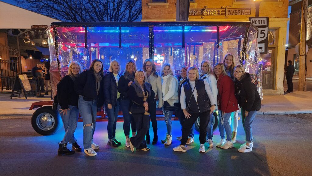 Group of people posing in front of a brightly lit trolley pub at night, wearing matching "Girl Trip in Progress" shirts.