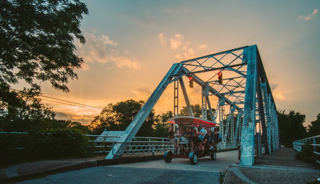 A pedal-powered vehicle with riders crosses a blue steel bridge at sunset.