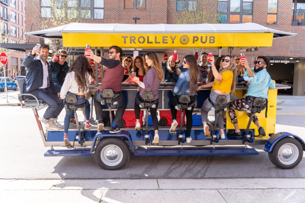 People on a pedal-powered trolley pub, toasting drinks on a sunny day.