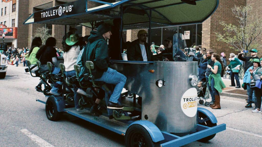 Pedal-powered "Trolley Pub" vehicle with passengers on a city street during a public event.