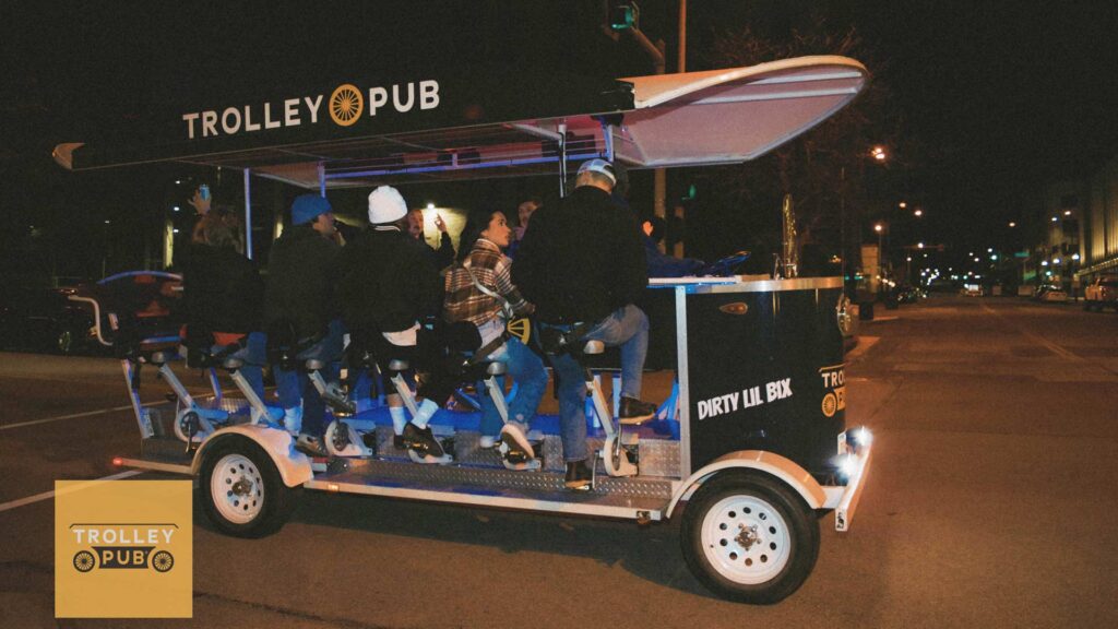 A group of people riding a multi-passenger, pedal-powered Trolley Pub at night on a city street.