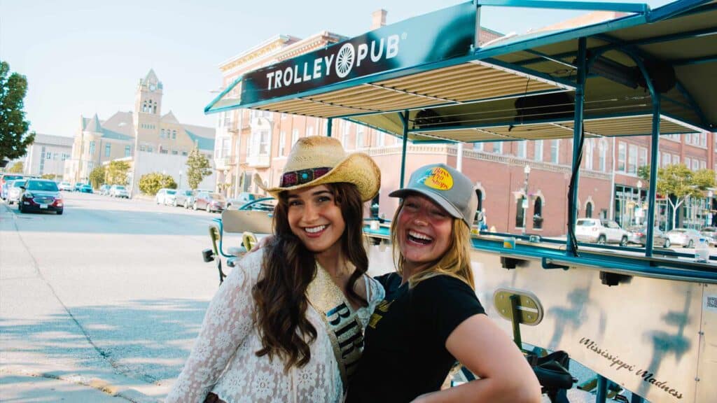 Two people near a Trolley Pub vehicle on a sunny street with buildings in the background.