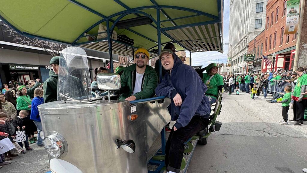 A pedal trolley with passengers on a street during a parade with spectators in green attire.