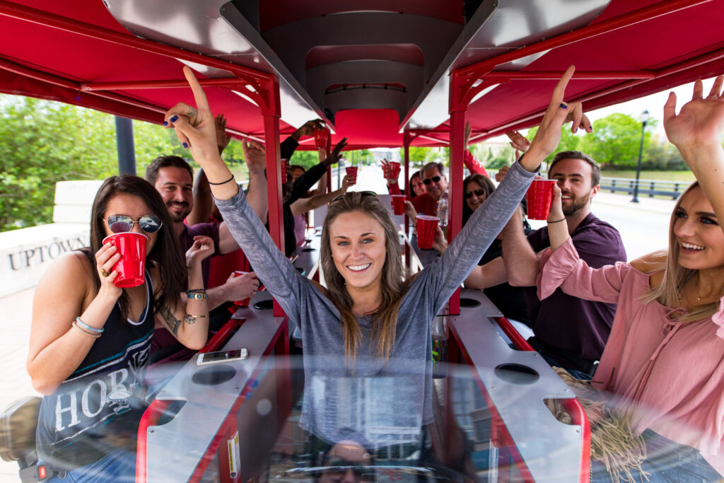 Group of people riding on a pedal pub, raising their arms and holding cups.