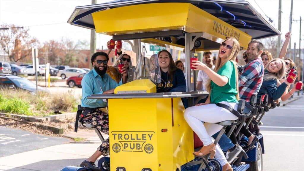 A group of people smiling and holding red cups while riding a yellow trolley pub bike on a sunny day.