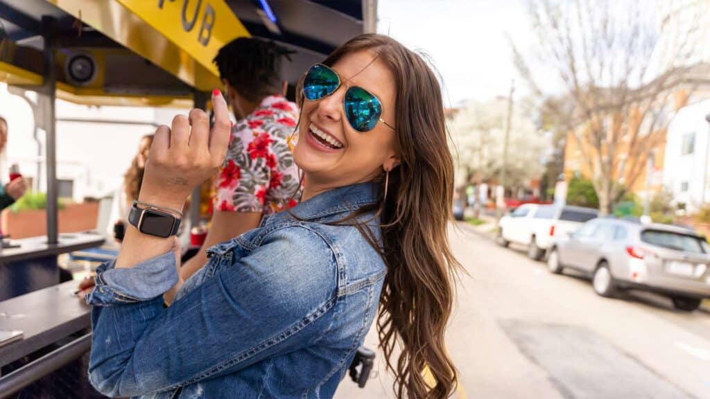 Woman wearing sunglasses and a denim jacket smiling outdoors with parked cars and trees in the background.